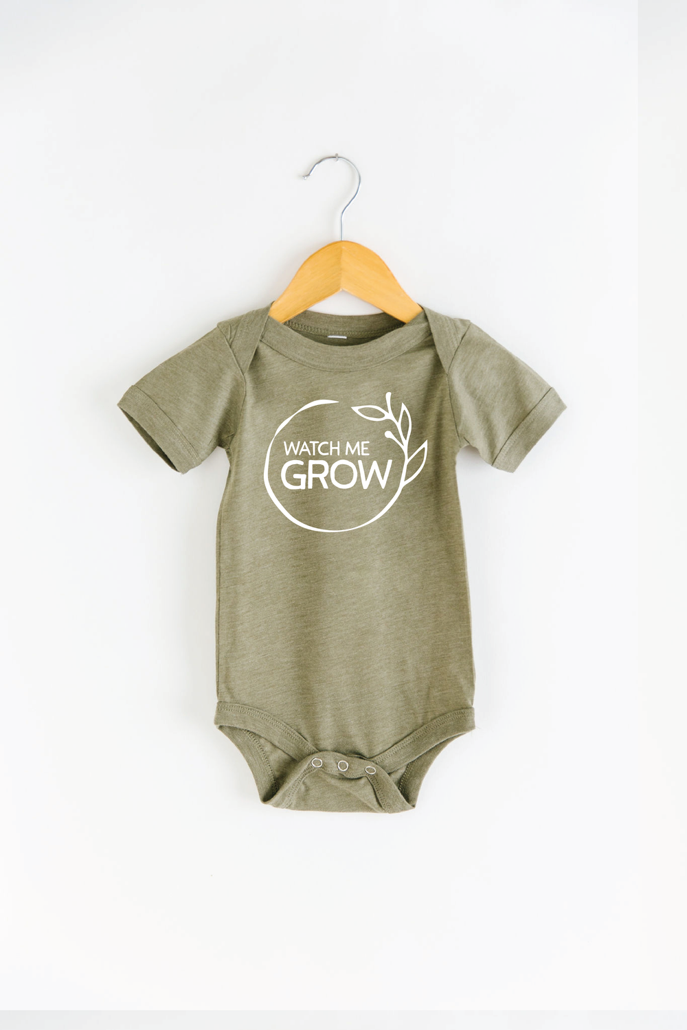 Olive green baby bodysuit on wooden hanger with white "Watch Me Grow" leaf design graphic