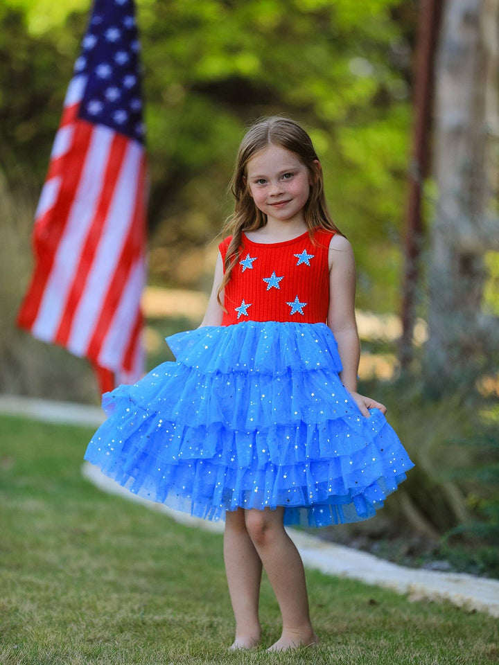 Young girl in a red and blue dress with star patterns standing outdoors with an American flag in the background.