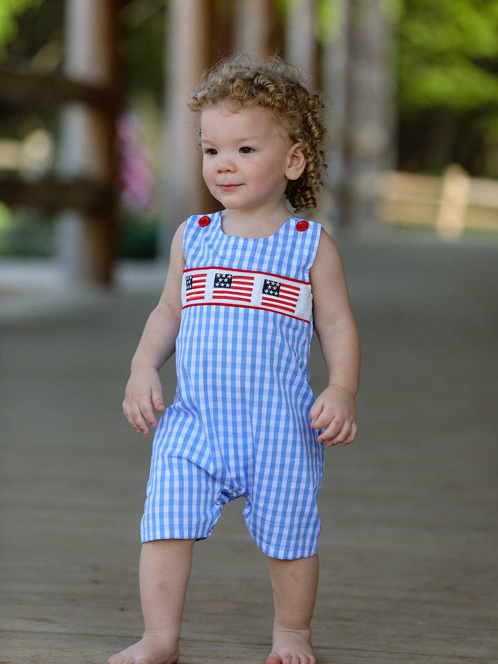 Child wearing a blue checkered romper with American flag design on a wooden deck.